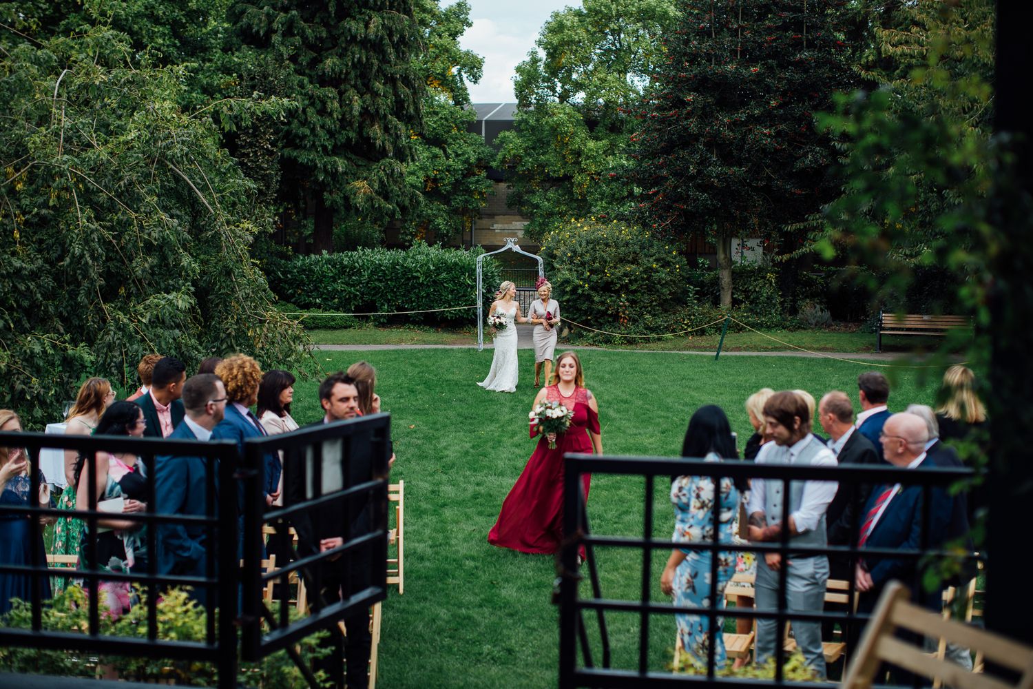 Outdoor wedding ceremony setup with guests seated on wooden chairs between black railings on a green lawn.