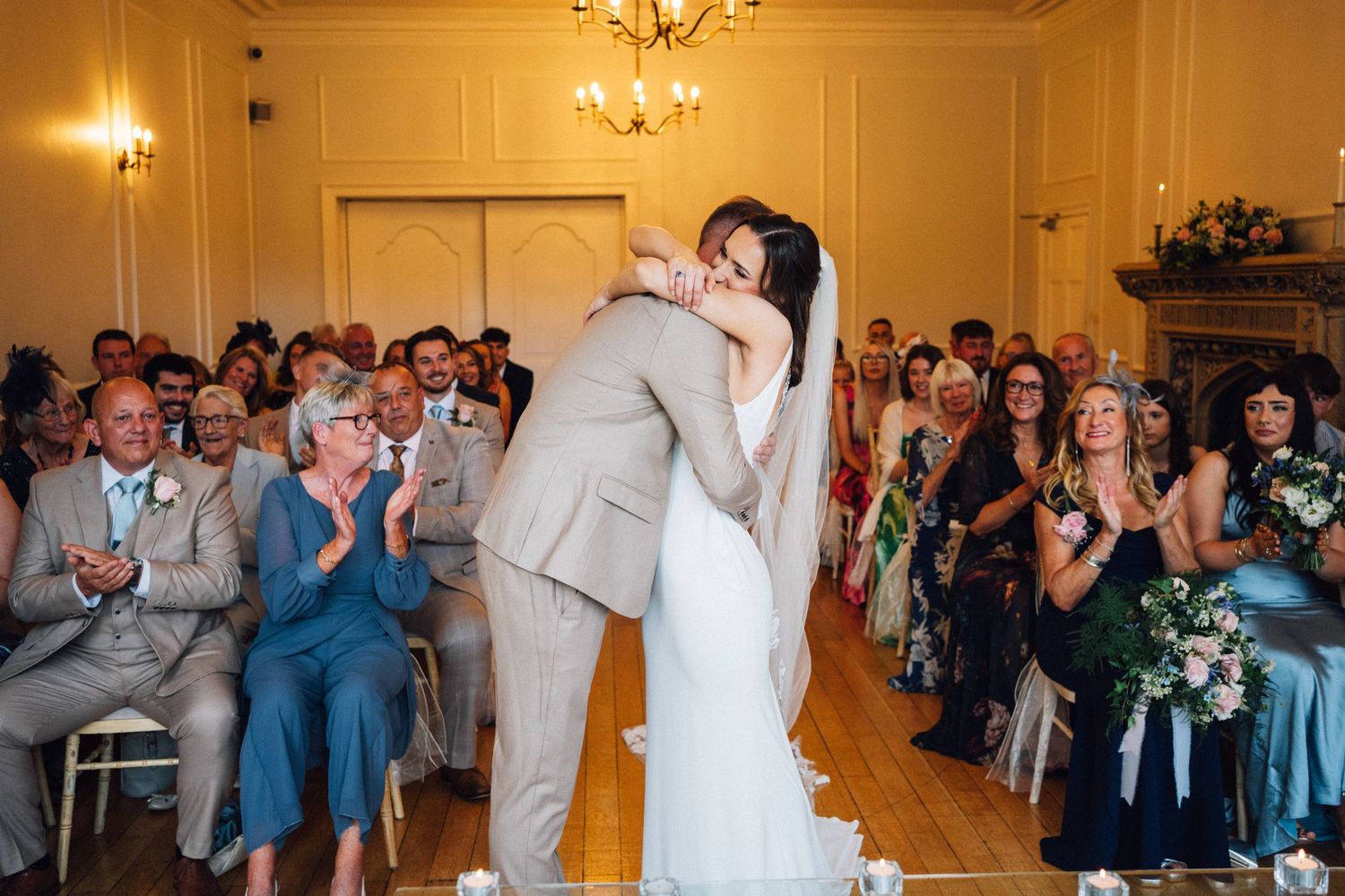 Wedding guests applaud during a ceremony in an elegant room with warm lighting and chandelier.