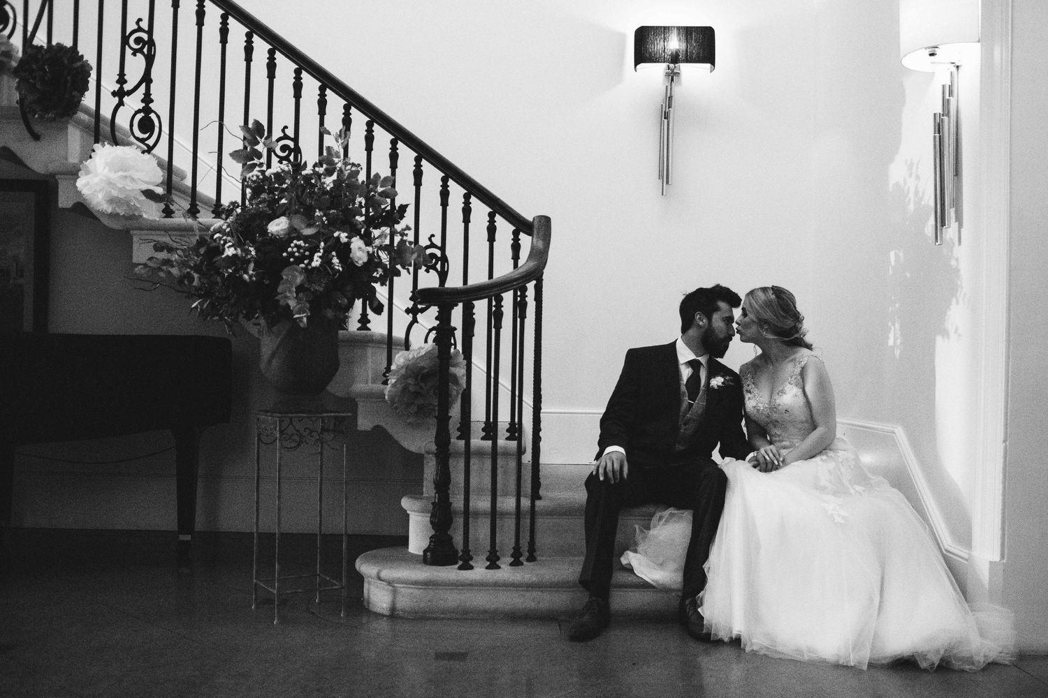 A couple in wedding attire descends an elegant staircase with wrought iron railings in a black and white photograph.