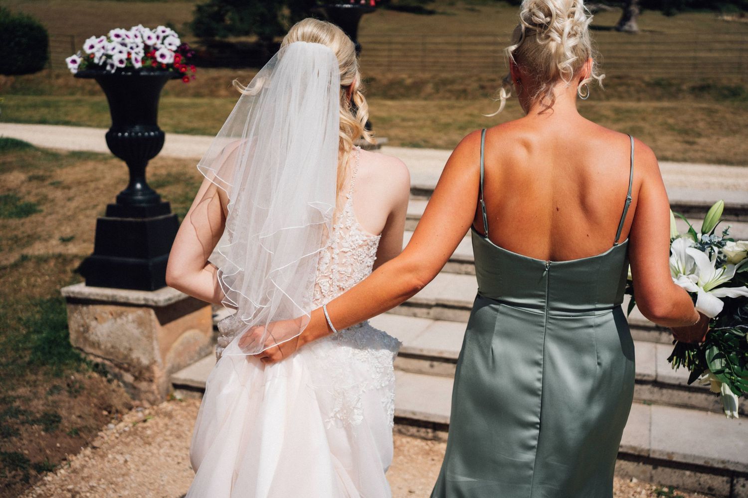 Two women in wedding attire walking together outdoors, one in white dress and veil, other in sage green dress.