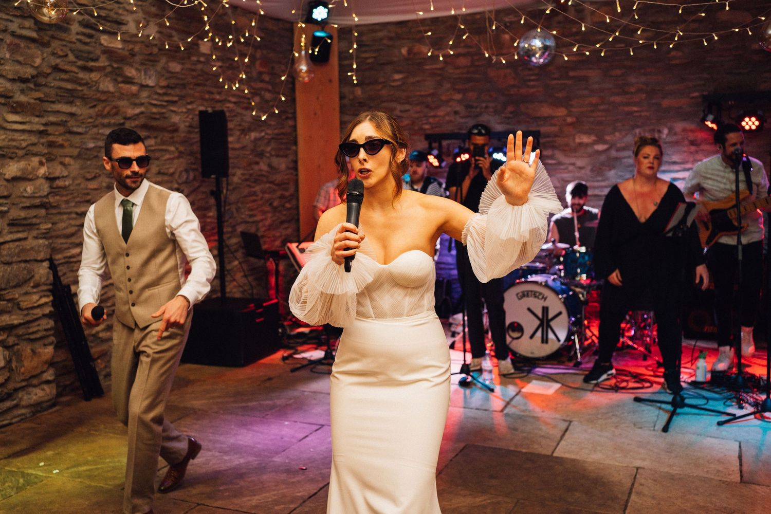 Wedding party dancing under string lights in rustic venue with stone walls.