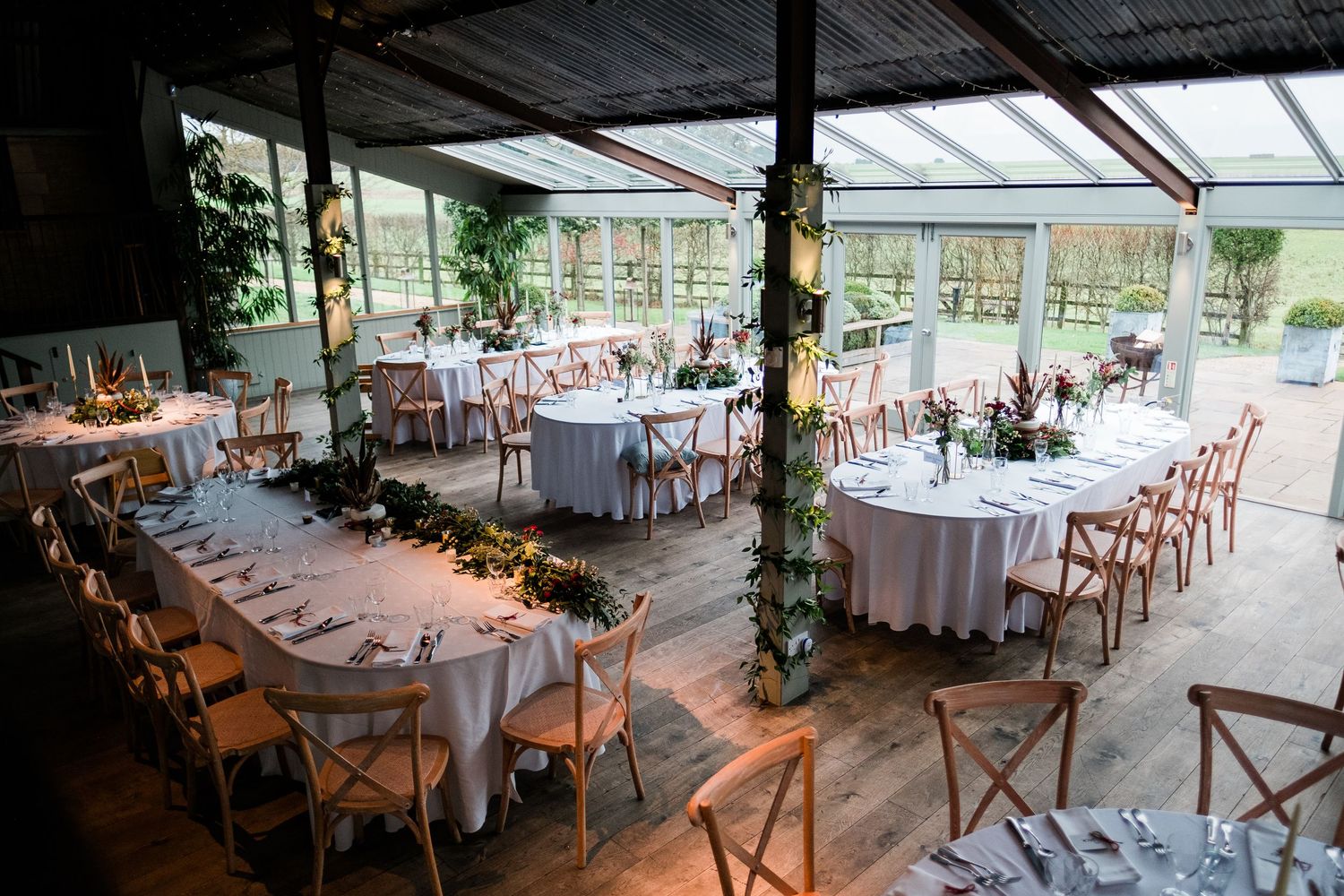 Reception hall set up with round tables and cross-back chairs under a glass ceiling.
