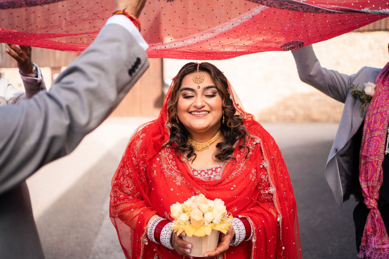 Smiling South Asian bride in red embroidered lehenga holding white flowers under red dupatta canopy at outdoor wedding.