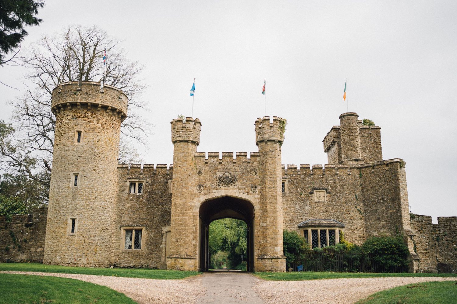 Medieval stone castle gatehouse with twin towers, arched entrance, and colorful flags under an overcast sky.