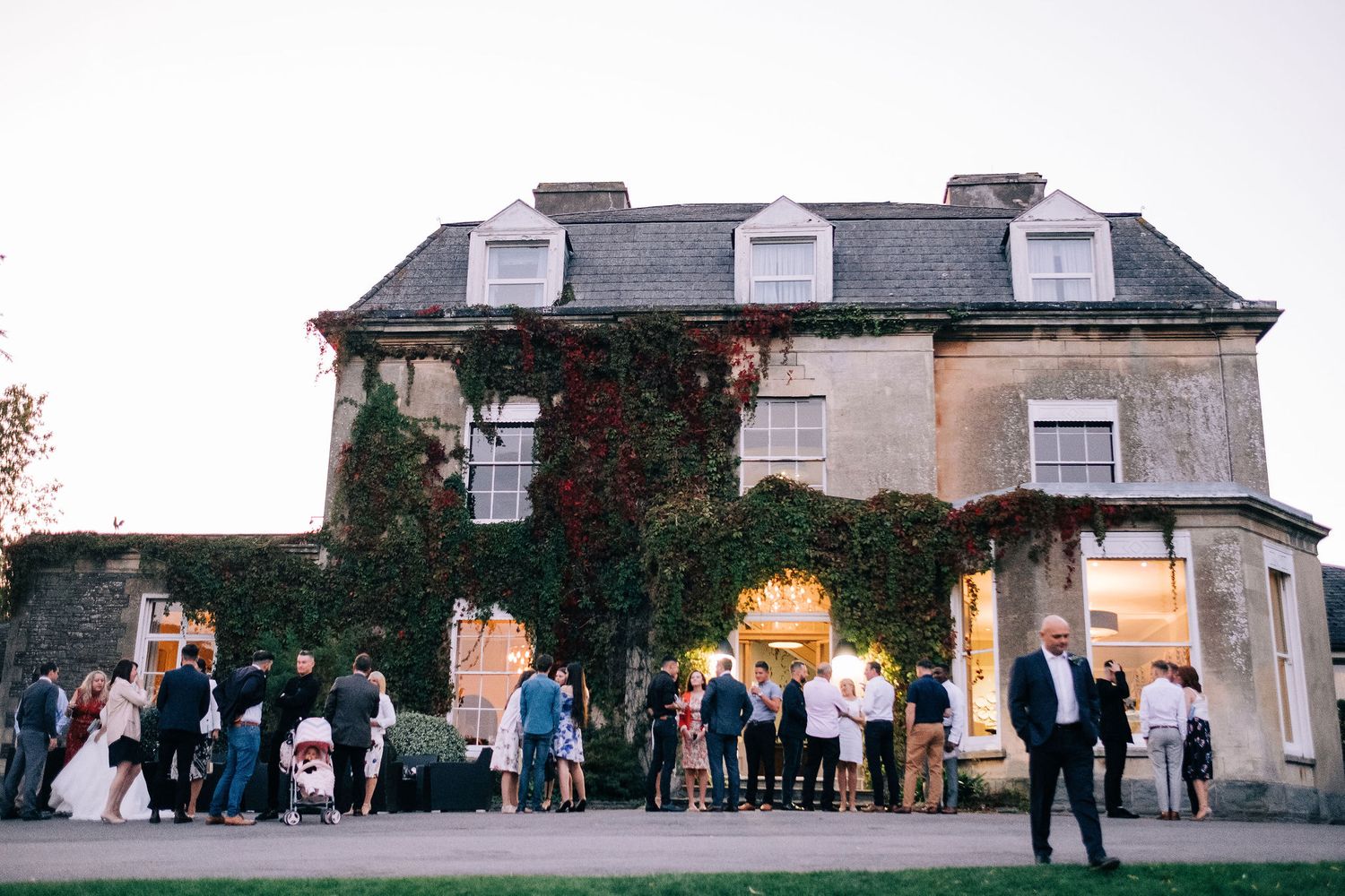 Wedding guests gather outside a stately venue at dusk during a celebration.