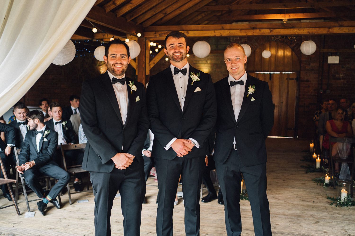 Groomsmen in formal black tuxedos stand together in a rustic barn setting with white paper lanterns.