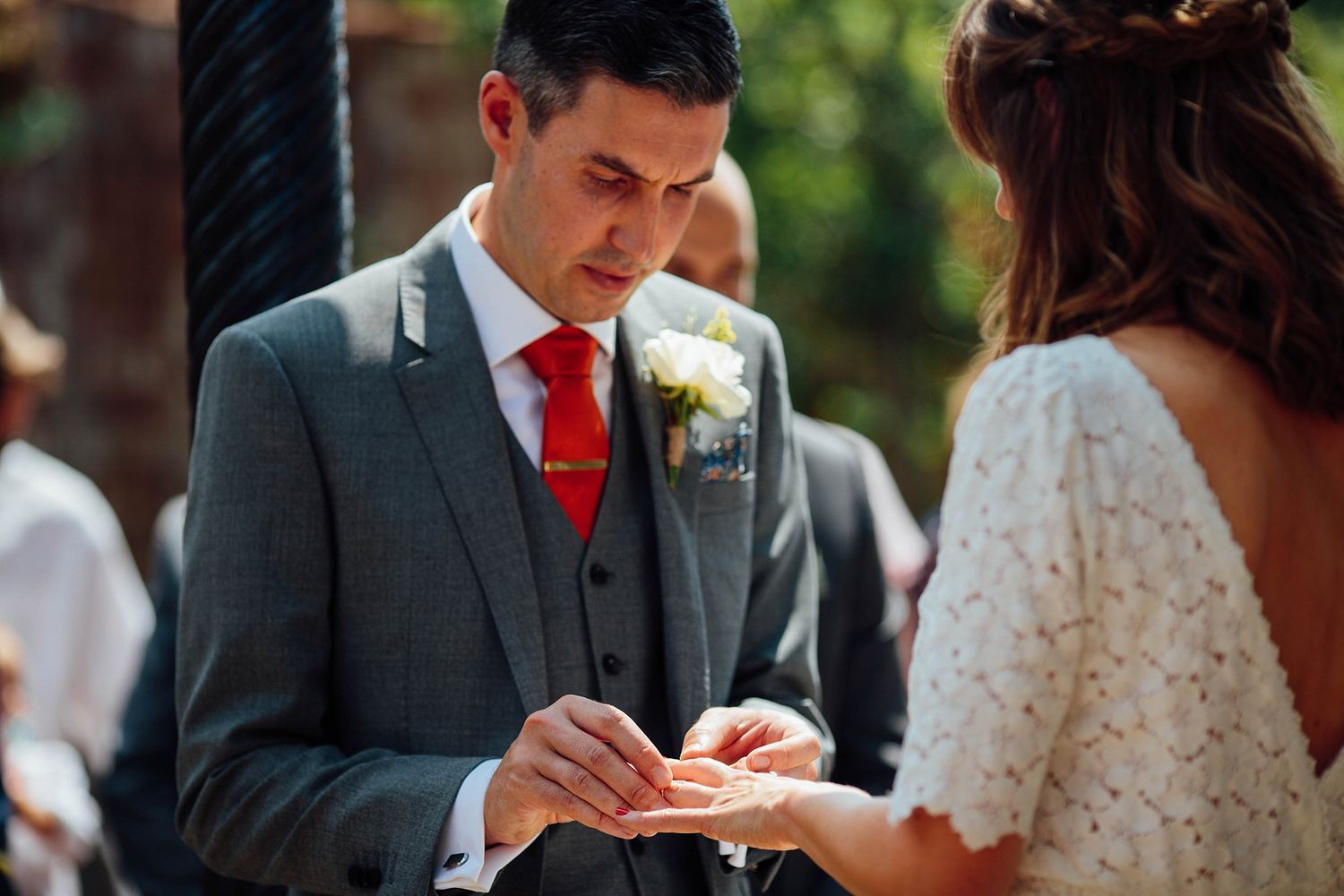 Wedding ceremony guests gather outdoors on a sunny day at a brick-walled venue with decorative columns.
