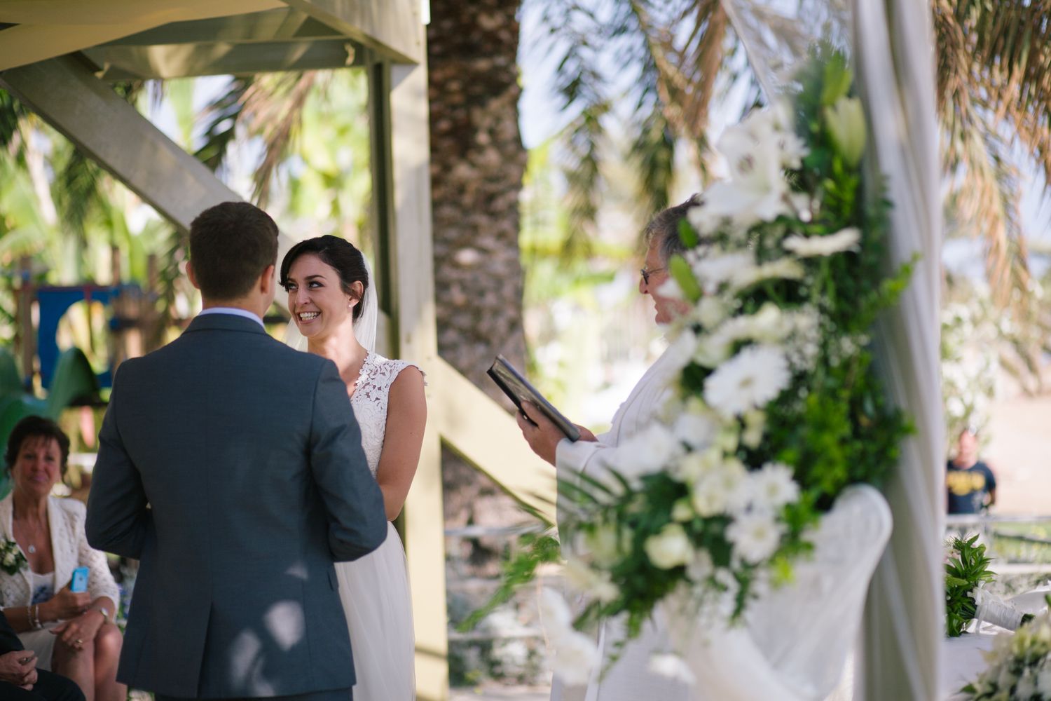 Outdoor wedding ceremony under palm trees with white decor and tropical setting.