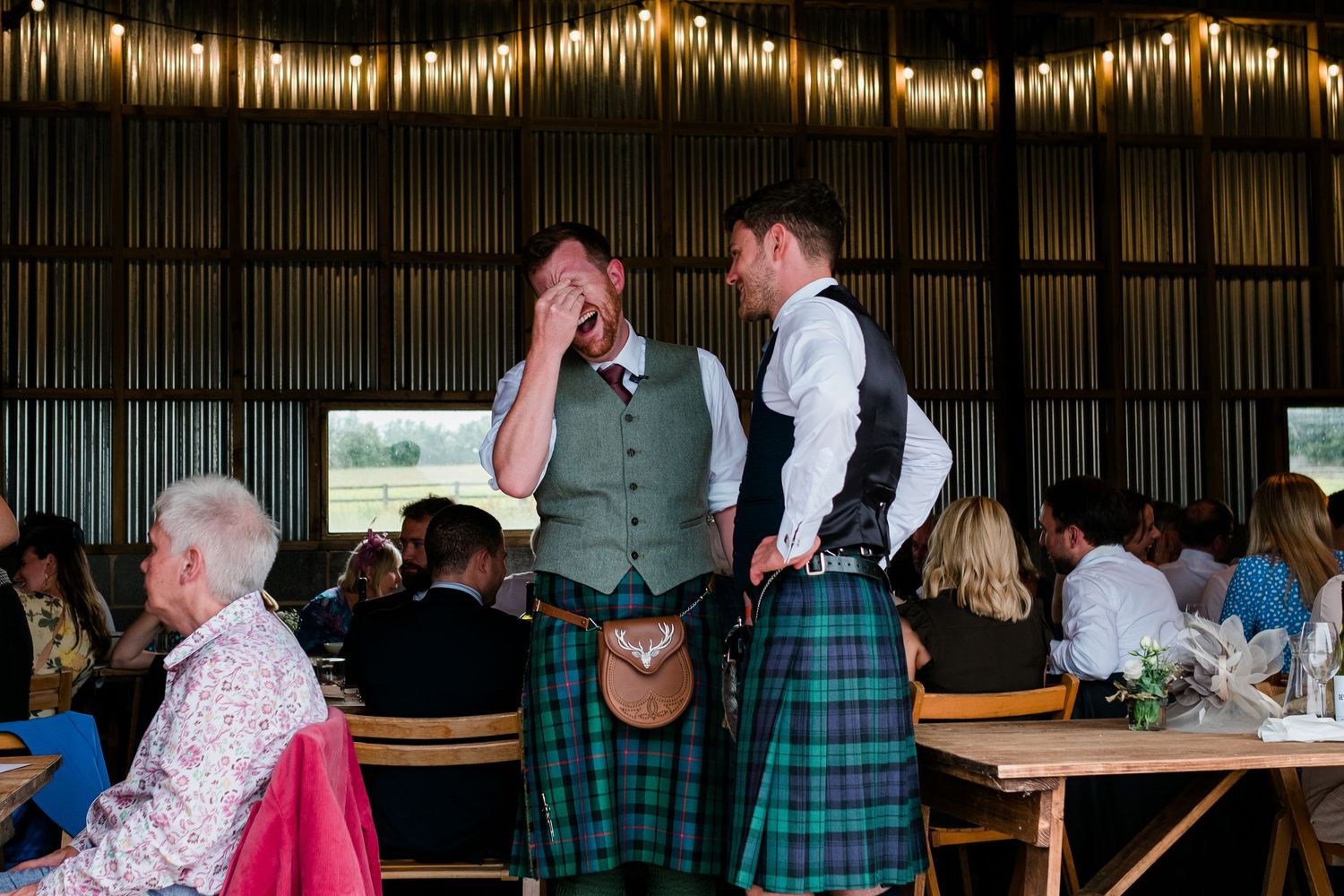 Two people wearing kilts and formal attire interact at an indoor wedding reception.