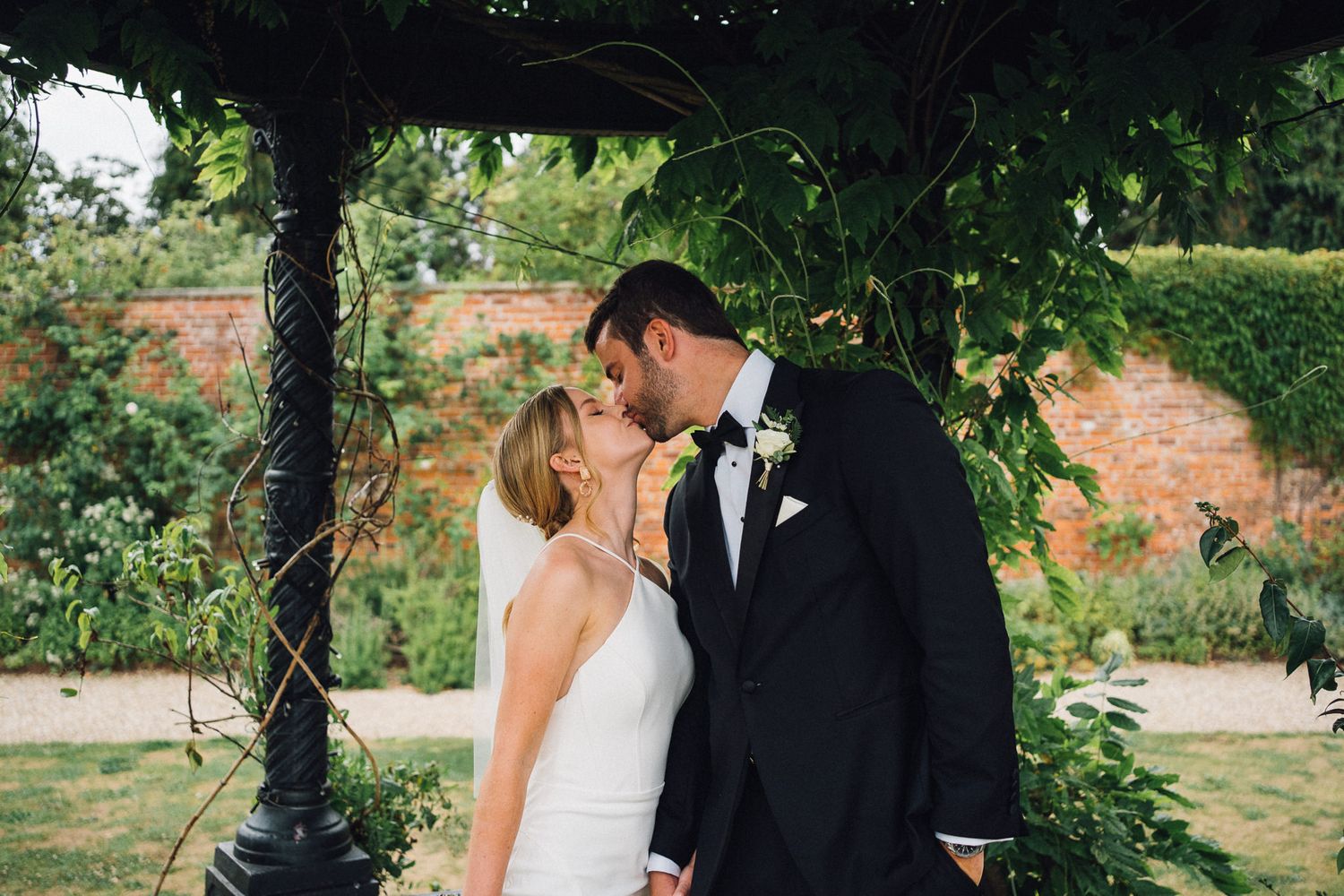 Romantic garden wedding portrait under an ornate black metal gazebo with lush greenery.