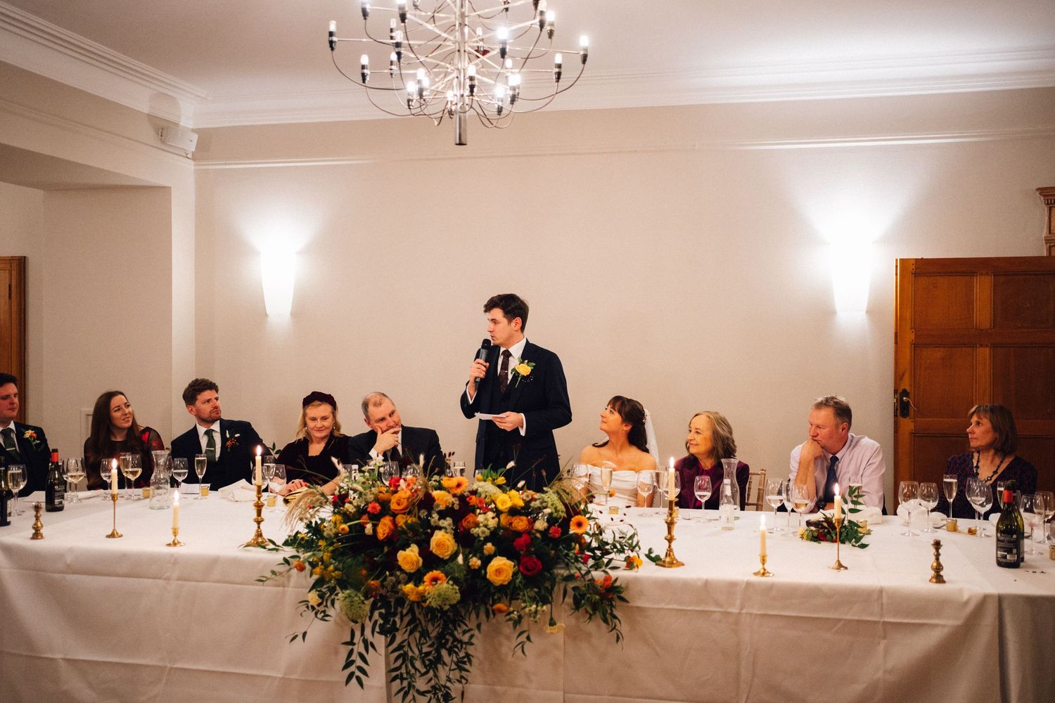 Wedding reception venue with elegant chandelier and long head table decorated with floral arrangements and candles.