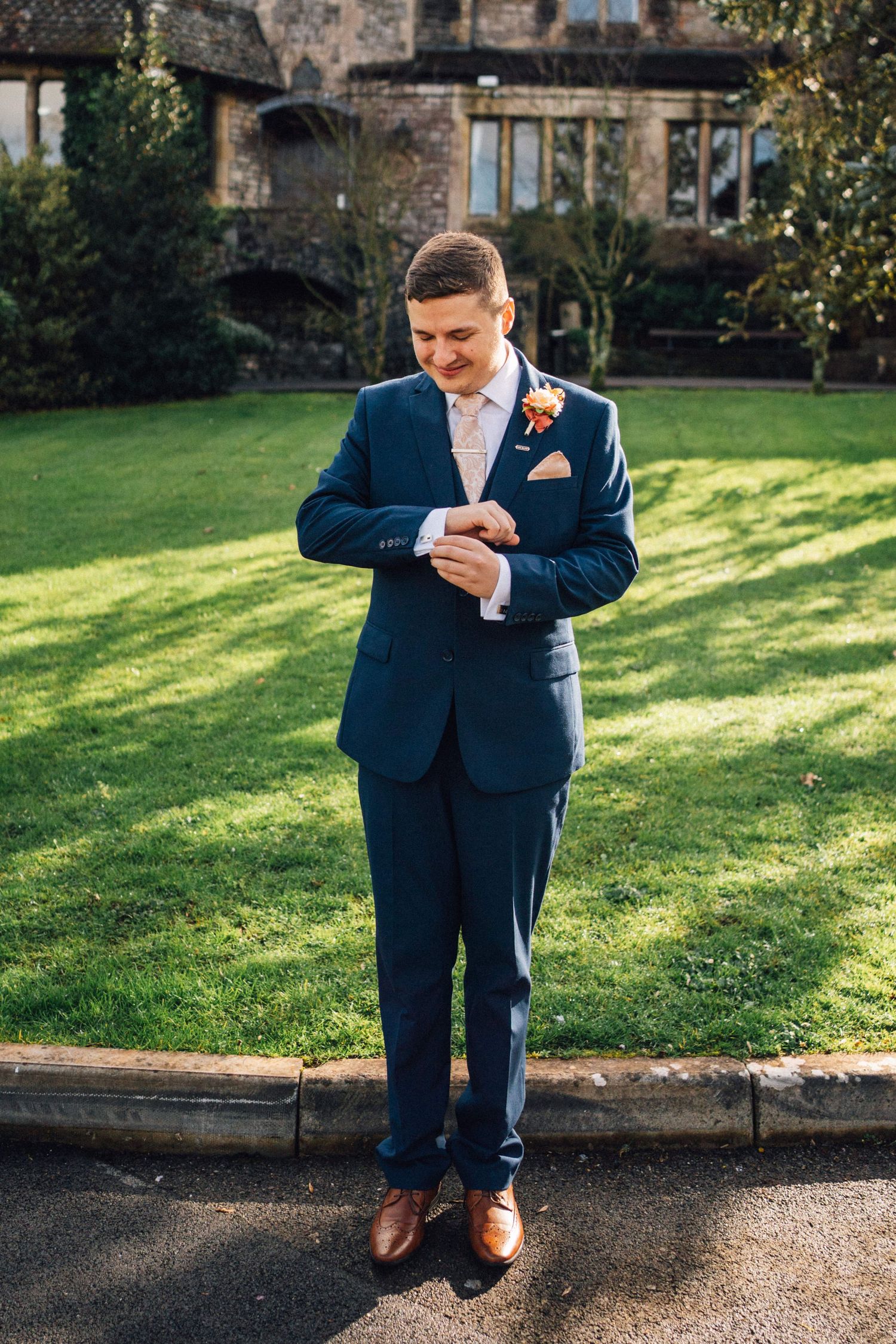 Person in navy blue suit checking their watch while standing on stone path near grassy lawn and historic building.