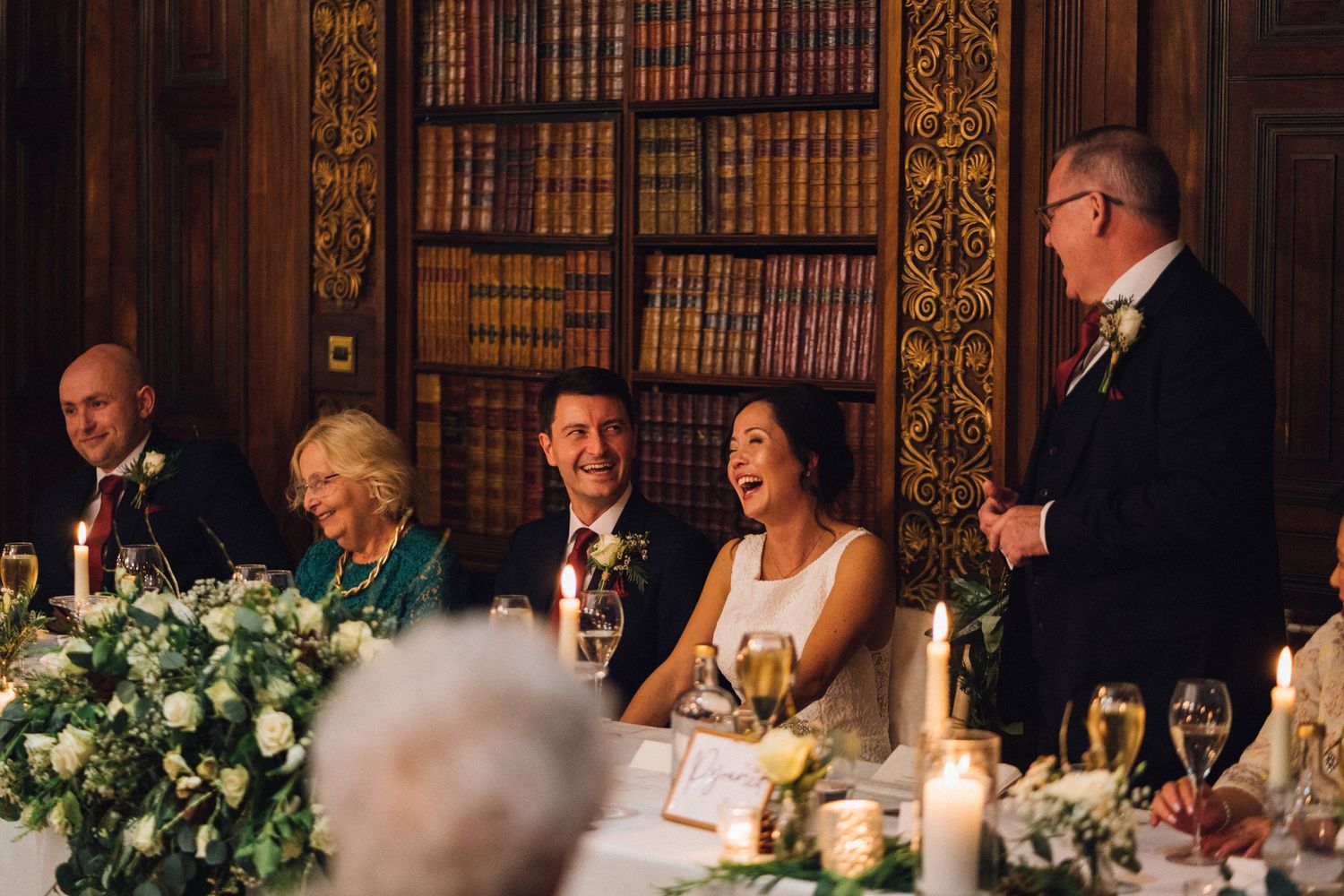 Wedding reception guests seated at an elegant candlelit table with floral centerpieces in a grand dining room.