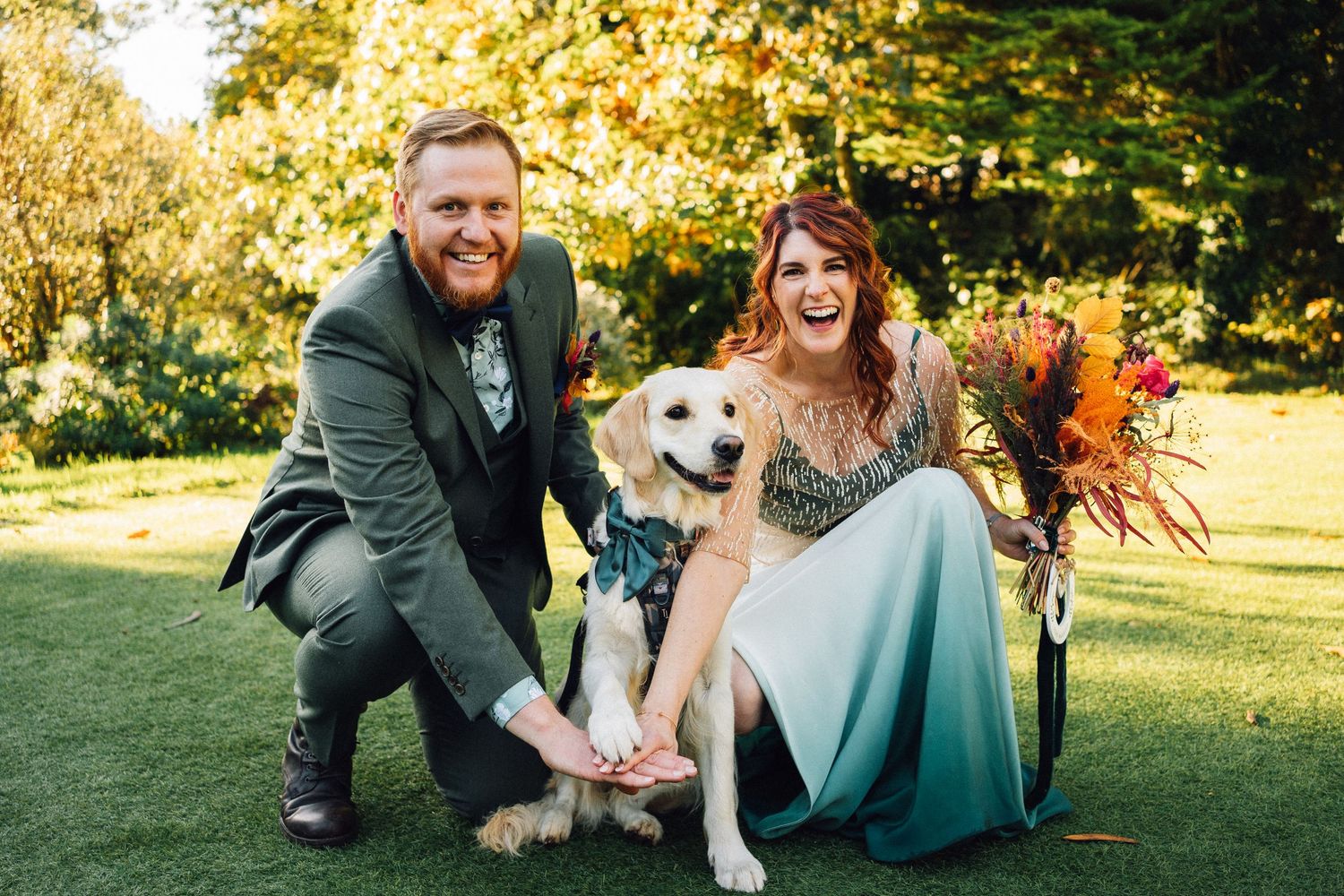 Wedding couple sits with their golden retriever on lush grass during sunset photo session.