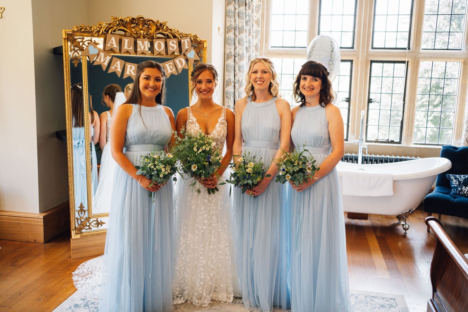 Bridesmaids in light blue dresses hold bouquets while standing in an elegant venue with large windows.