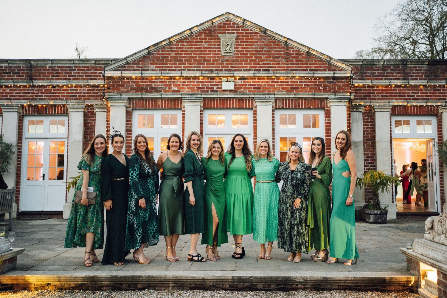 Wedding party in coordinated green dresses poses in front of brick building exterior.