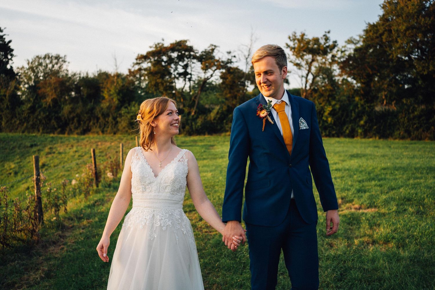 A couple walks hand in hand through a sunlit meadow during golden hour in their wedding attire.