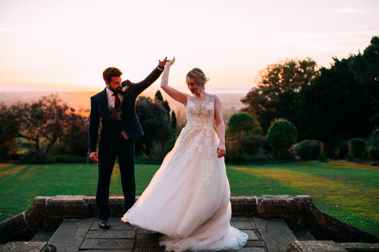 Bride and groom dance together at sunset on a scenic hilltop with rolling landscape views in the background.