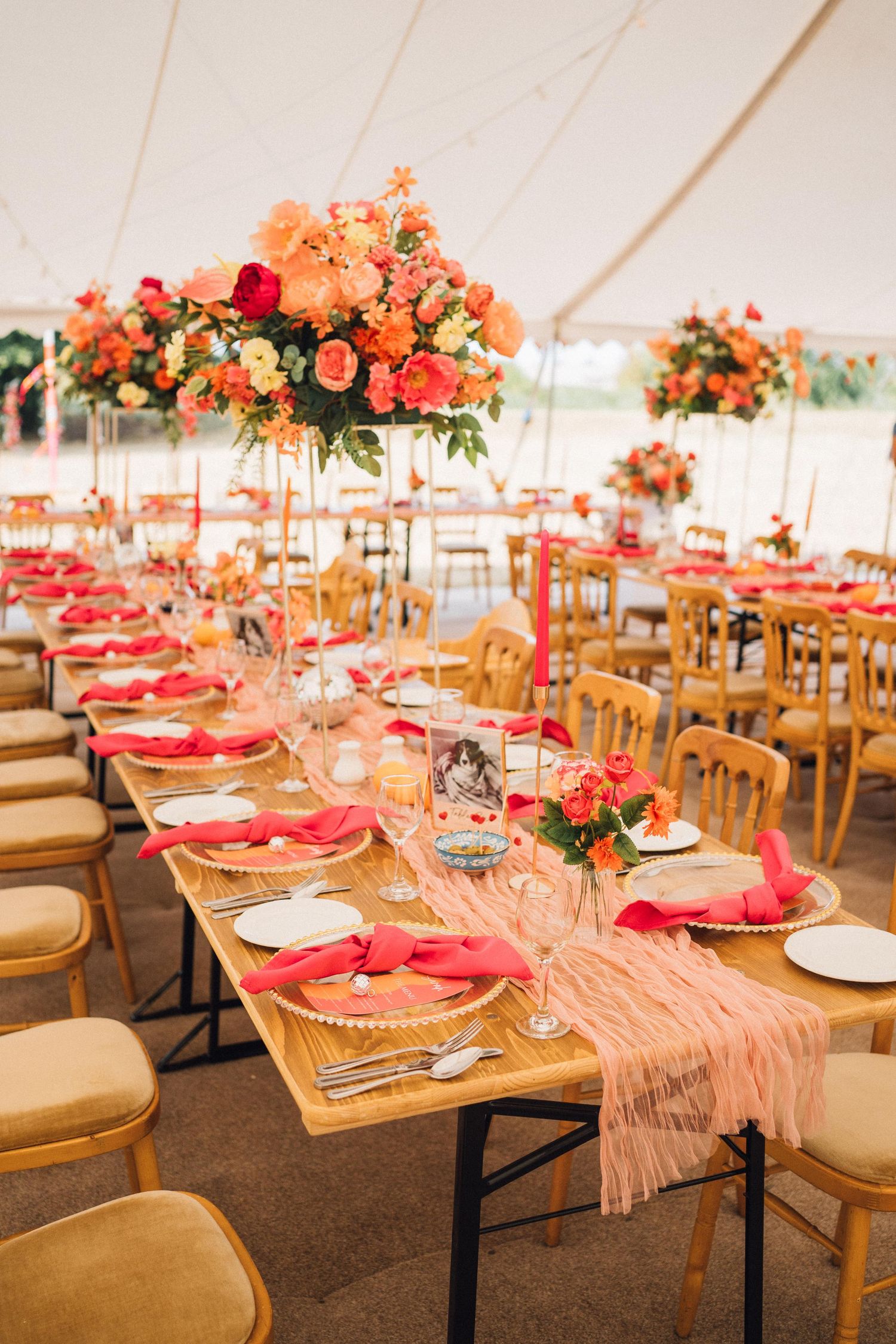 Elegant wedding reception table setting with coral napkins, tall floral centerpieces and wooden chairs under white tent.
