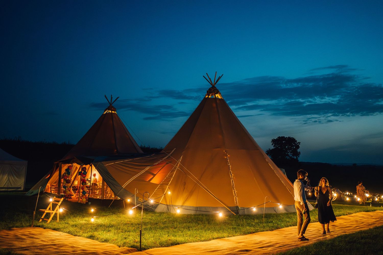 Illuminated traditional teepee structures at dusk create a magical camping atmosphere against the darkening sky.
