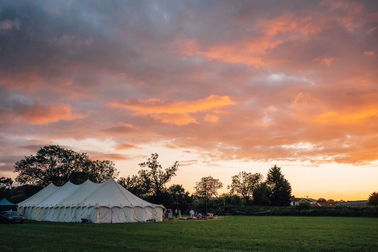 A white wedding tent stands in a grassy field under a dramatic orange and purple sunset sky.