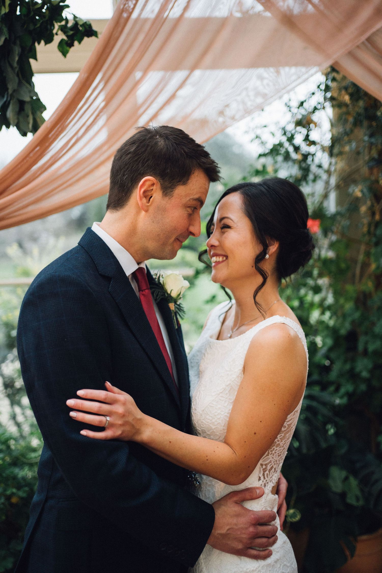 A romantic wedding couple embraces under a peach-colored canopy with lush greenery in the background.