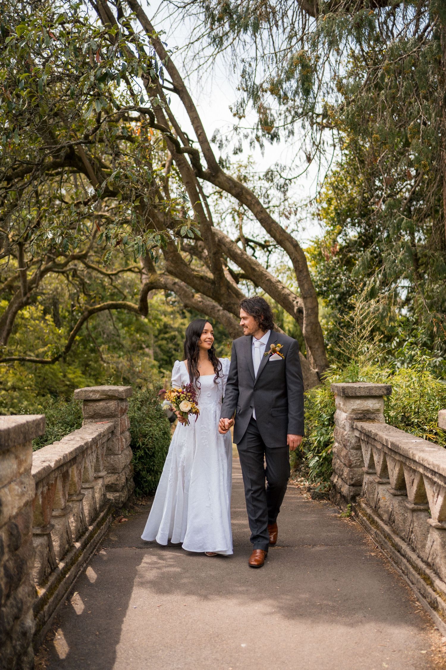 Bride and groom walk hand-in-hand on a stone bridge surrounded by lush trees, bride holding a colorful bouquet.