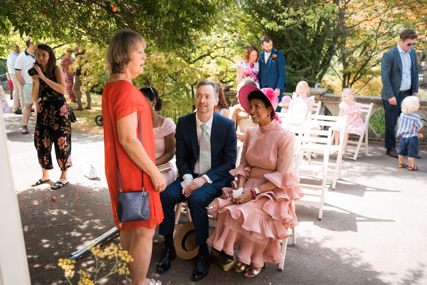 Guests socializing at an outdoor wedding ceremony with white chairs and lush green trees in the background.