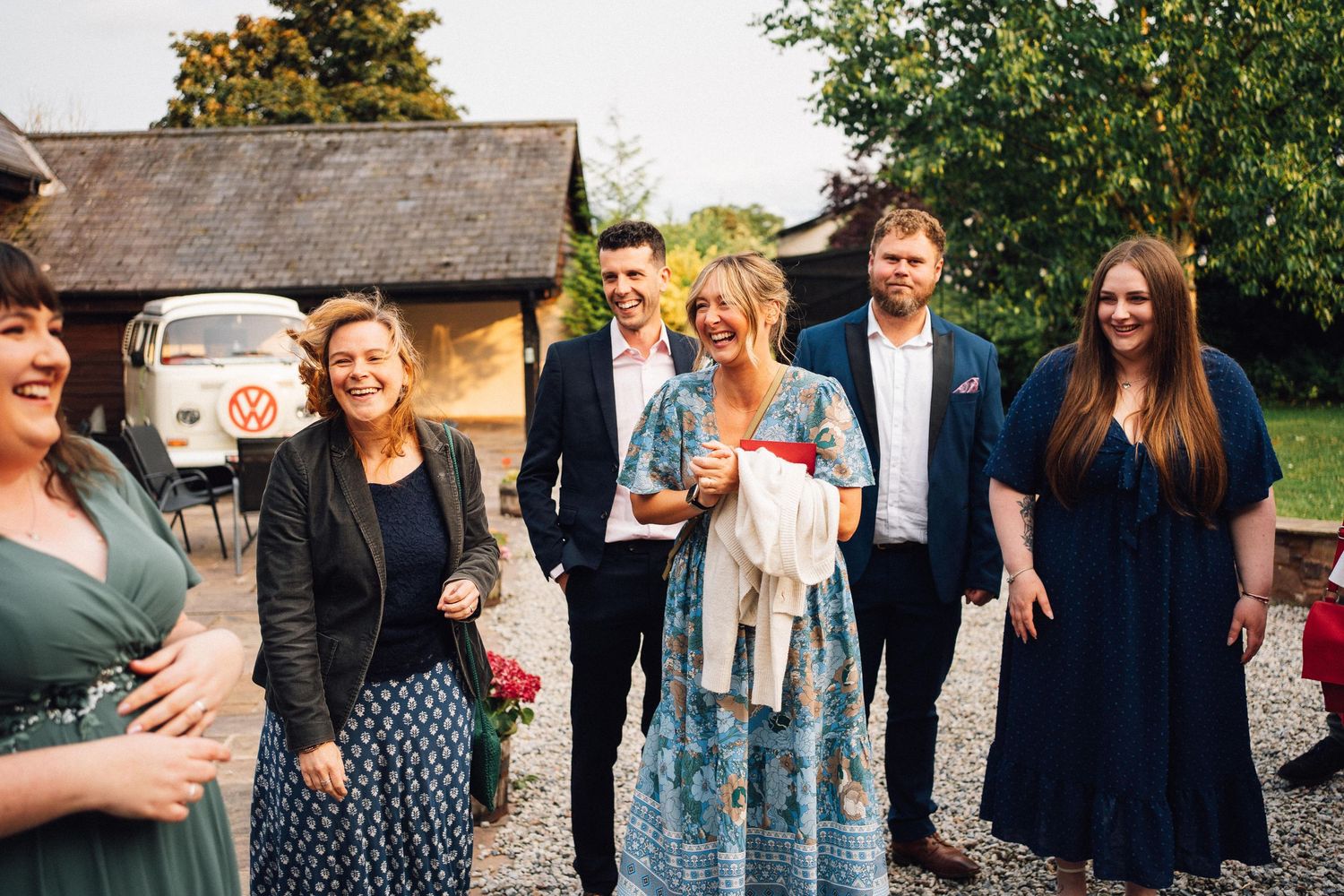 A group of people gathered outdoors for a social event in a garden setting with trees and greenery in the background.