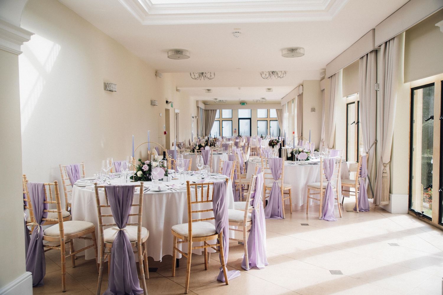Panoramic view of a bright white wedding reception room with lavender-accented chairs and round tables.