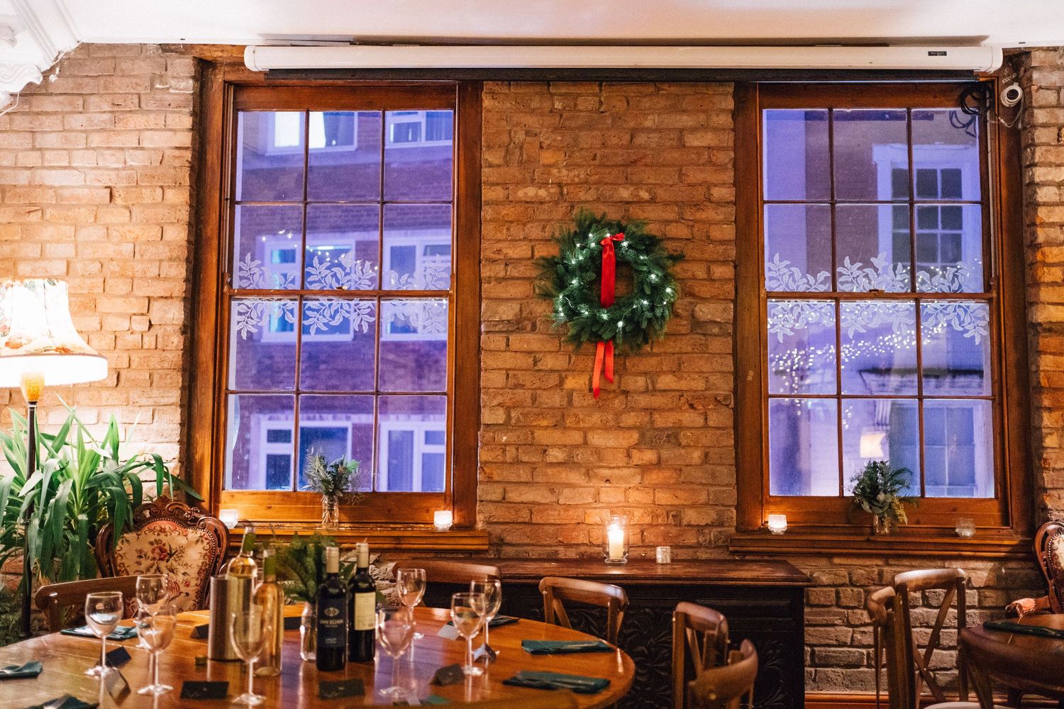 Cozy brick-walled restaurant interior with warm lighting, string lights, and holiday wreaths creating an intimate atmosphere.