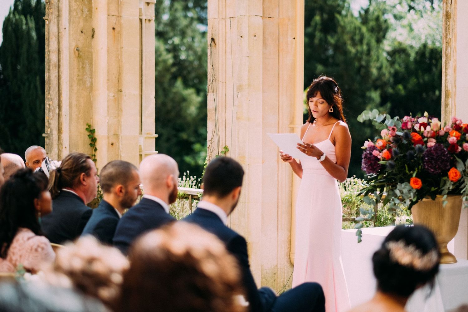 Wedding guests stand in a line at an outdoor ceremony venue with elegant stone columns and floral decorations.