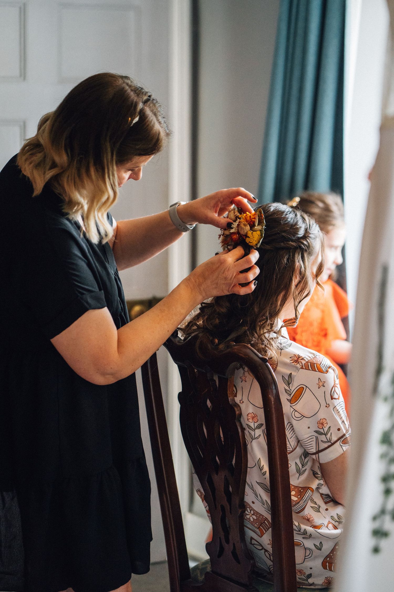A hairstylist works on styling a client's long dark hair in a salon setting with turquoise curtains in the background.