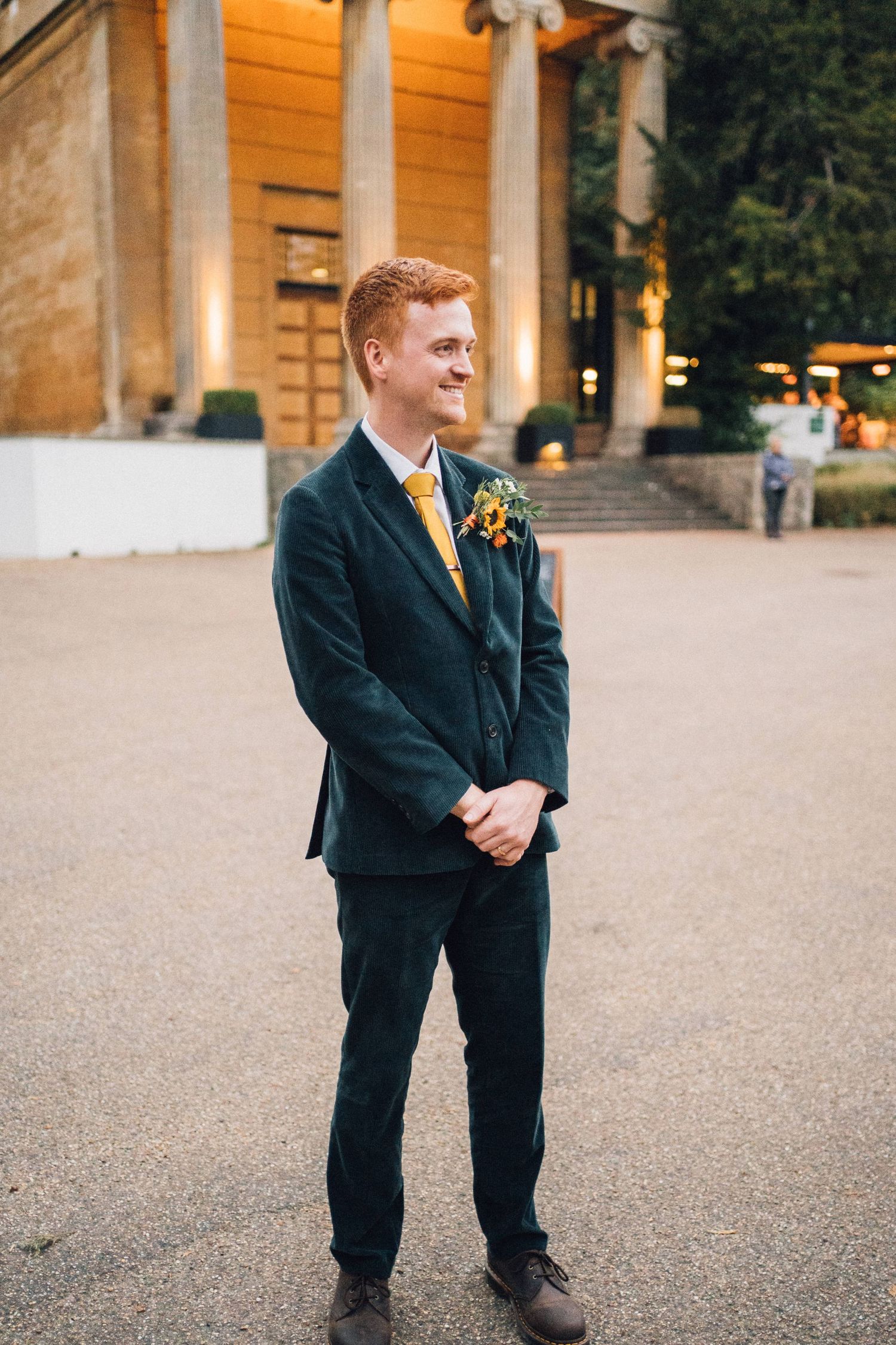A young ring bearer in a dark suit stands on a gravel path outside an elegant venue at dusk.