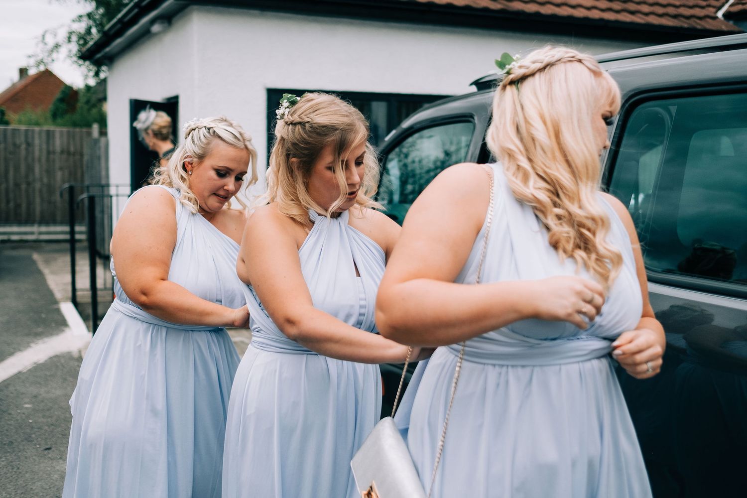 Wedding party members in light blue dresses embracing outdoors near parked cars.