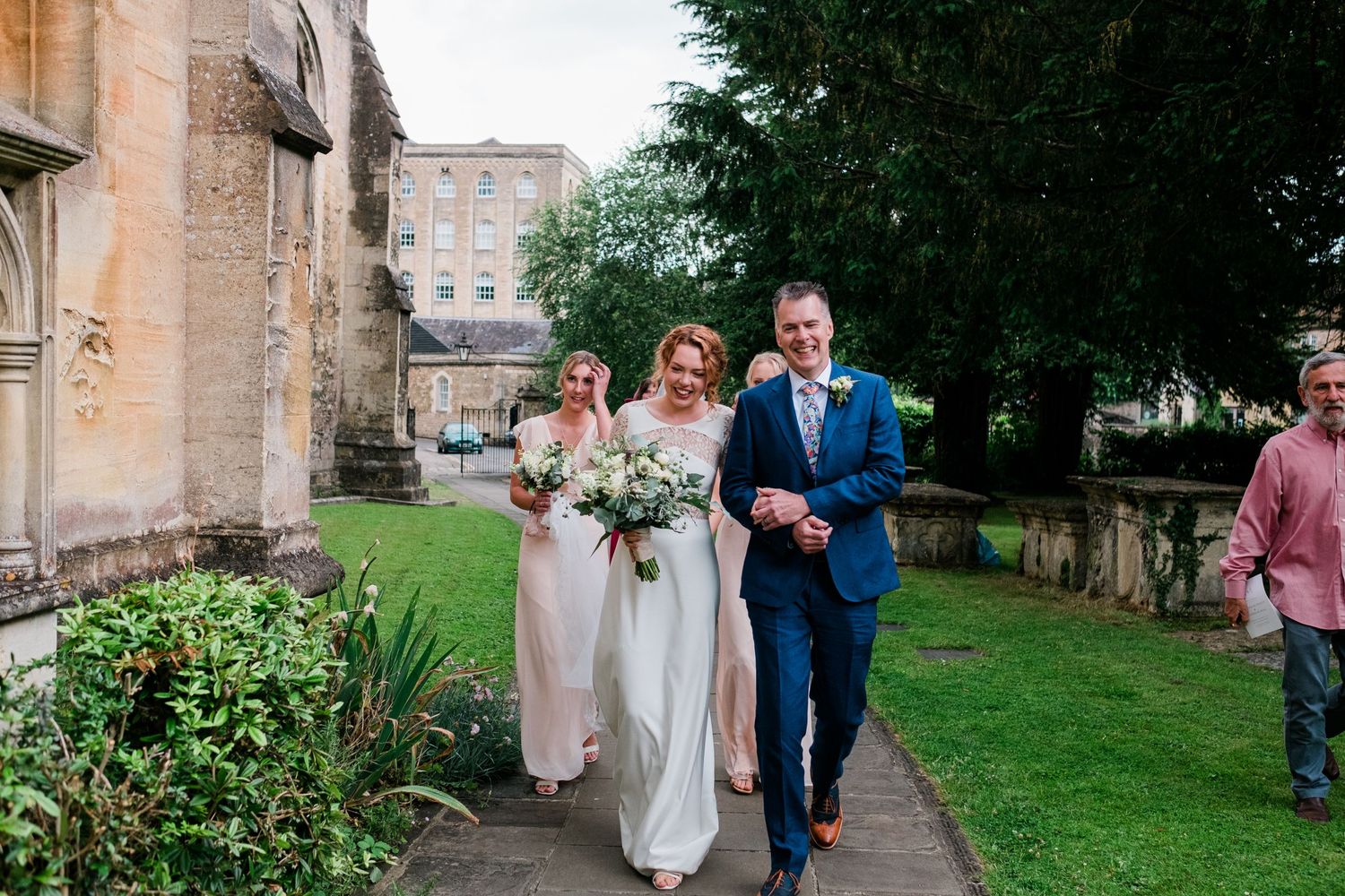 Wedding party walking through lush garden path beside historic stone church.