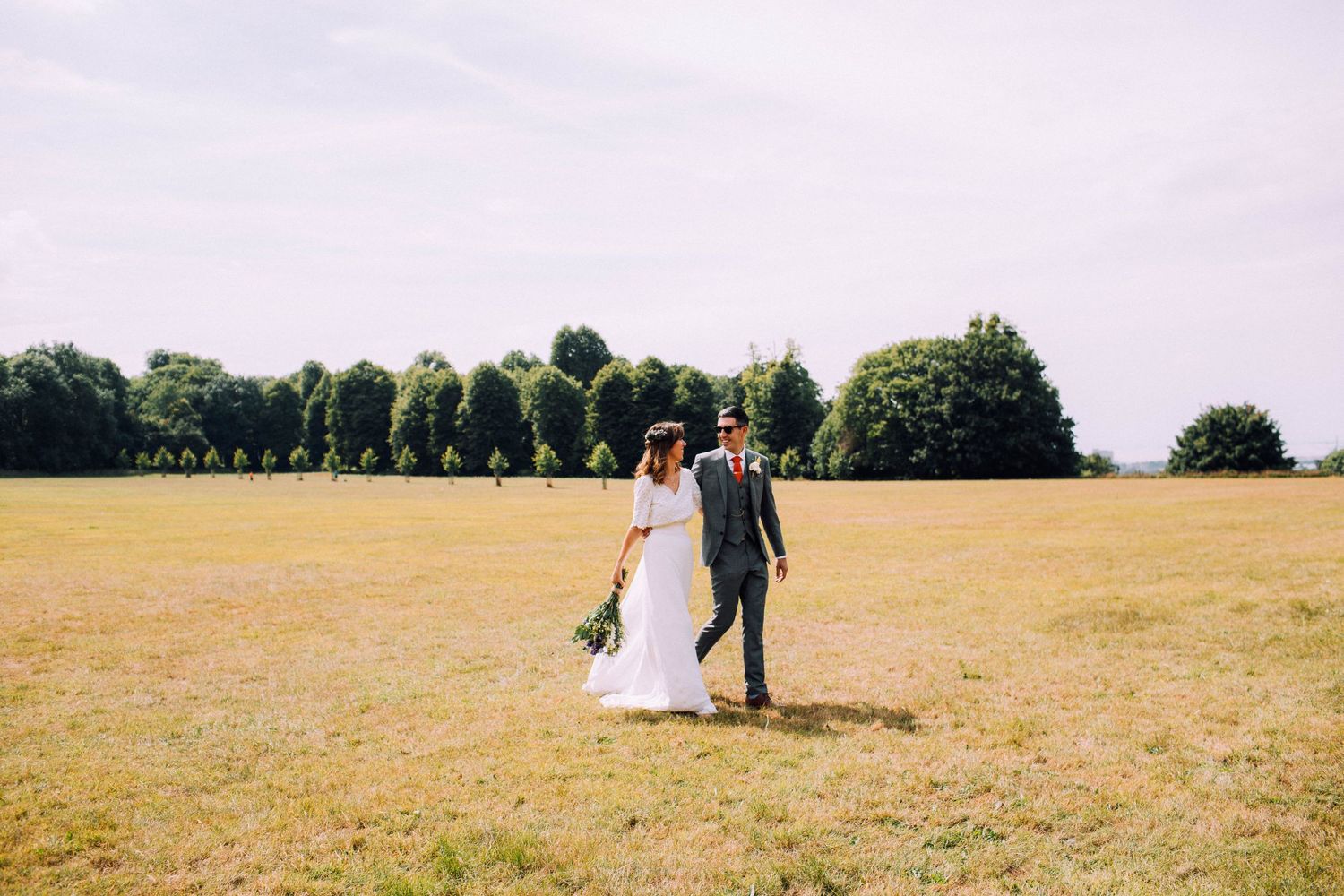 Couple walking through a sunny meadow on their wedding day.