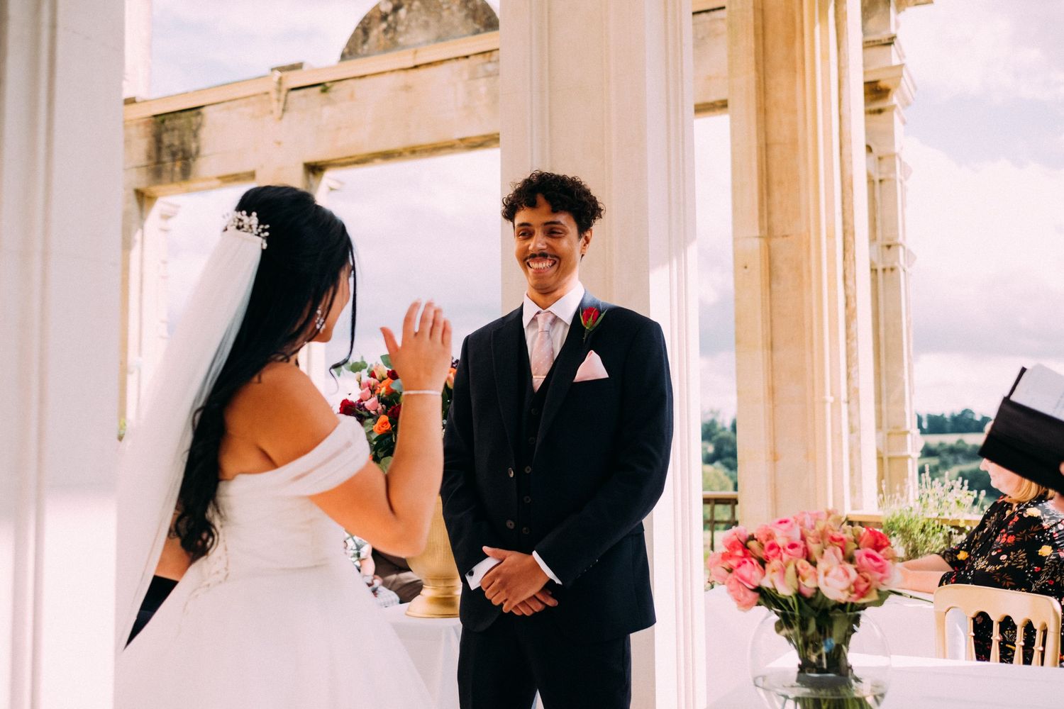 A couple exchanges vows during their wedding ceremony under elegant white columns with pink flowers.