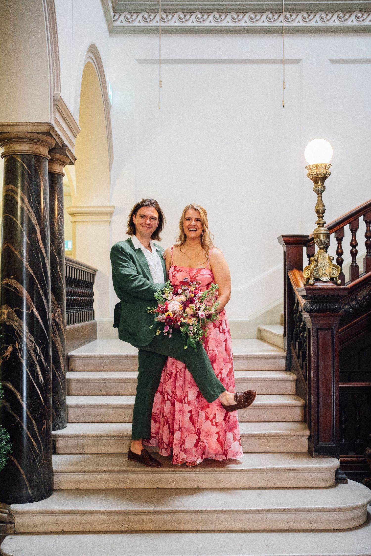 Couple posing on elegant white marble stairs with ornate wooden banisters.