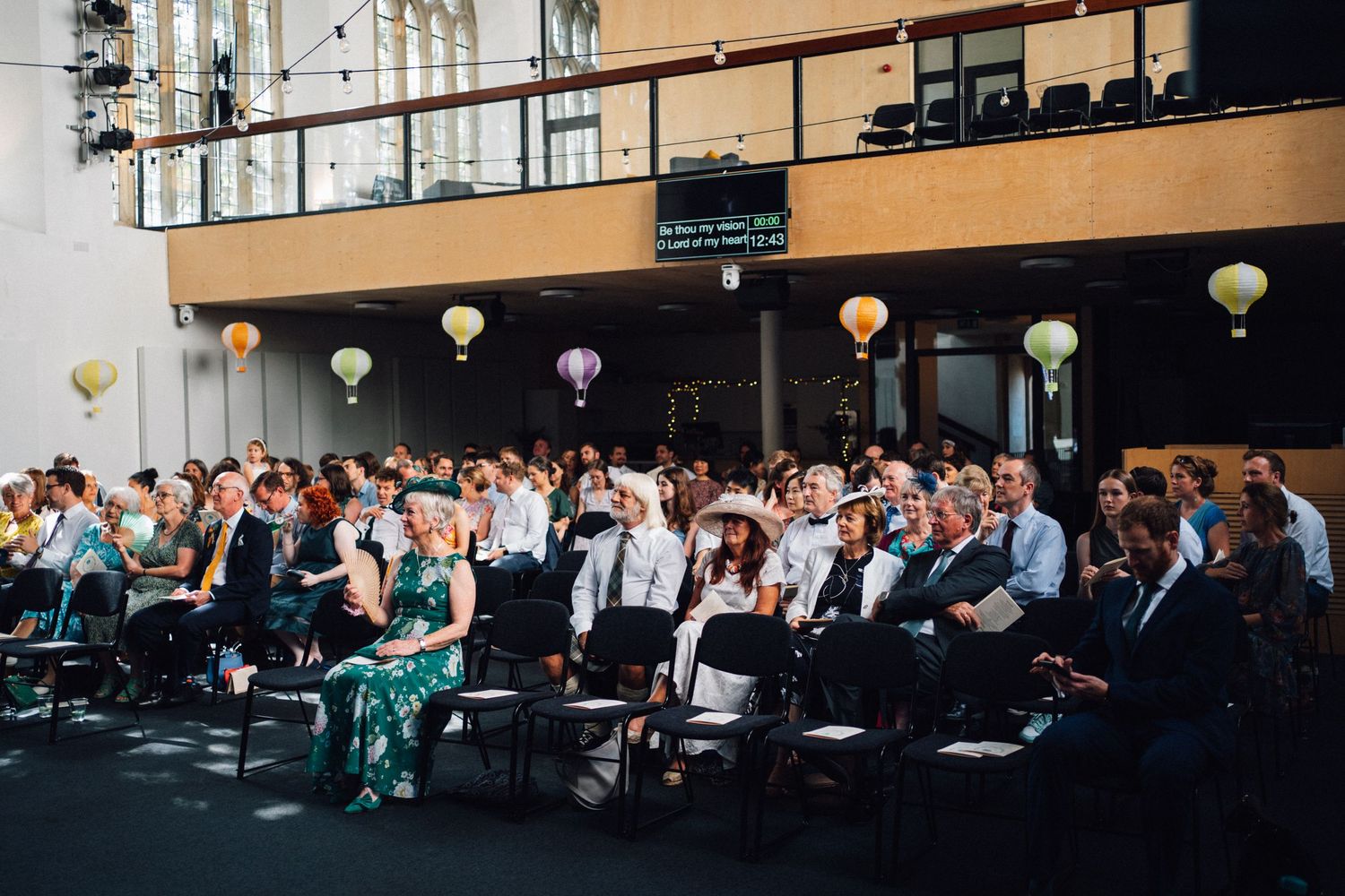 Wedding guests seated in rows at an elegant indoor ceremony venue with warm lighting and white chairs.