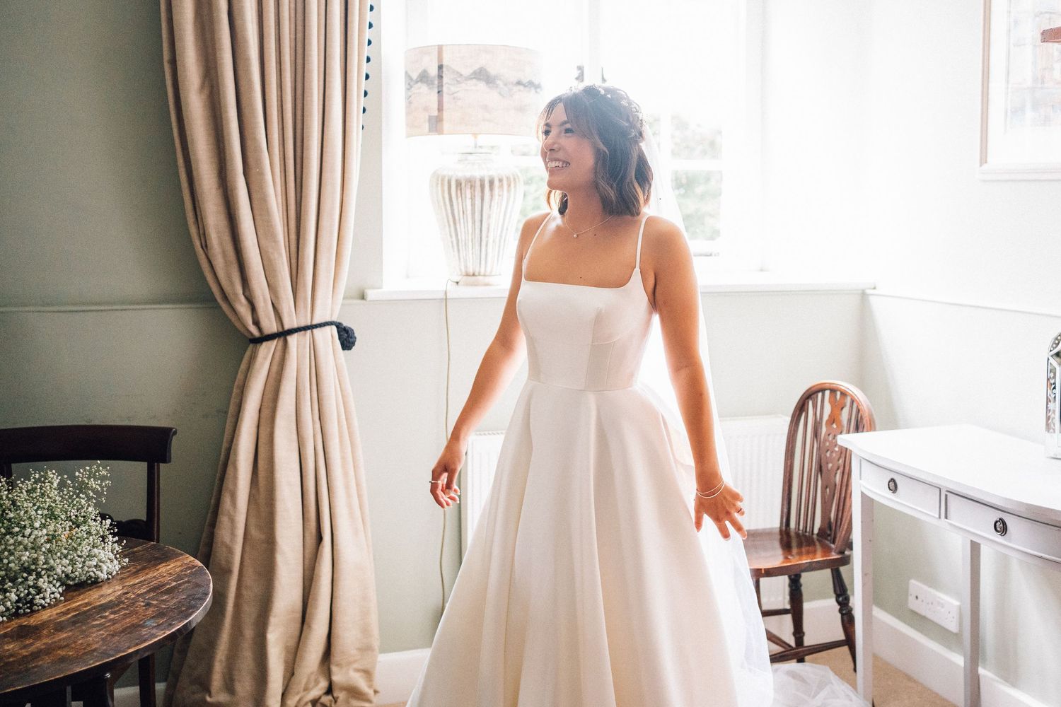 A bride in a classic white wedding gown stands by a window in an elegant room with beige curtains.