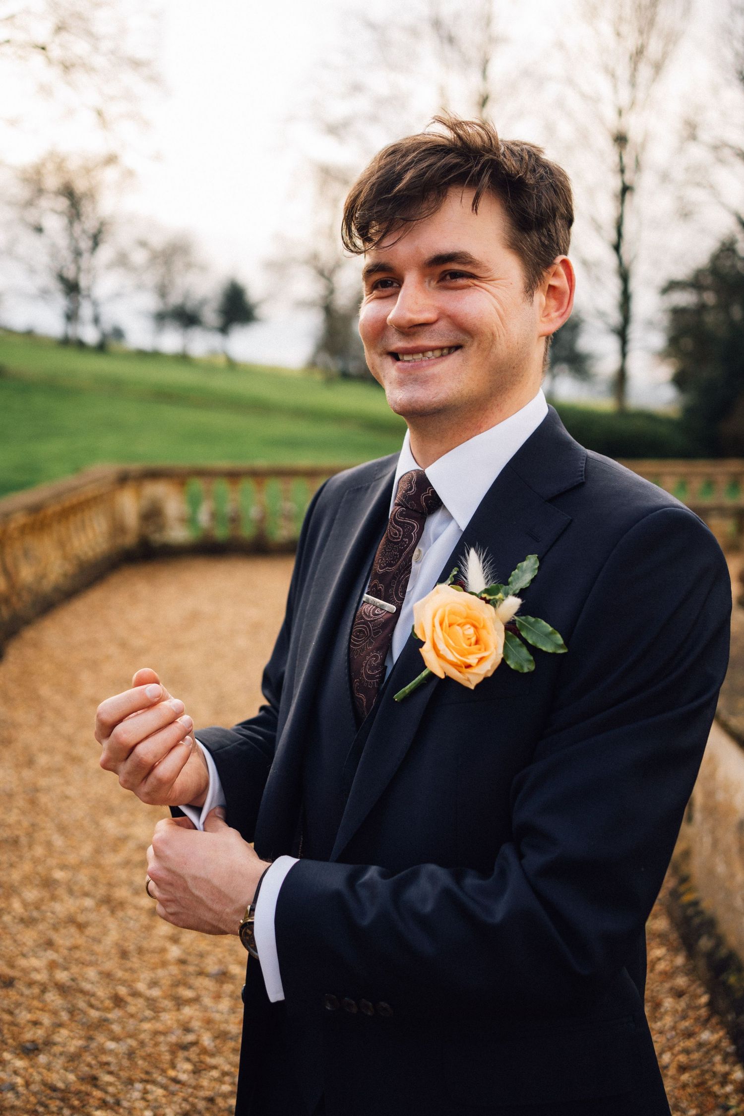 A person in a dark suit with an orange boutonniere smiles while standing in an autumn outdoor setting.