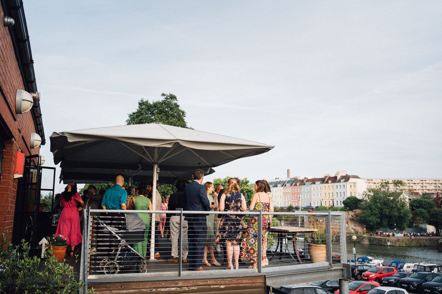 People socializing on a rooftop terrace bar with large umbrella overlooking colorful waterfront buildings on a cloudy day.