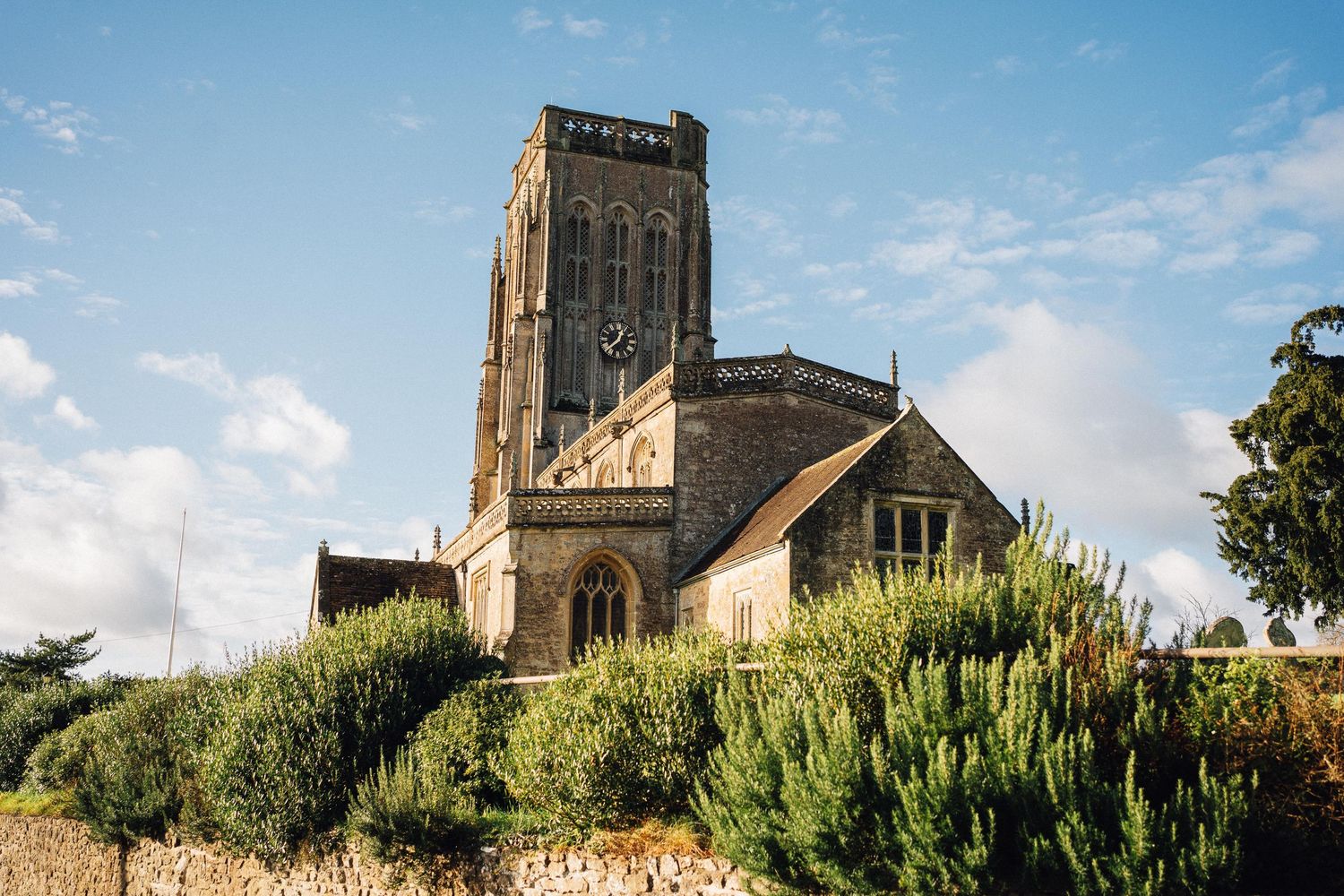 Historic stone church tower rises above trees and buildings against a bright blue sky in panoramic view.