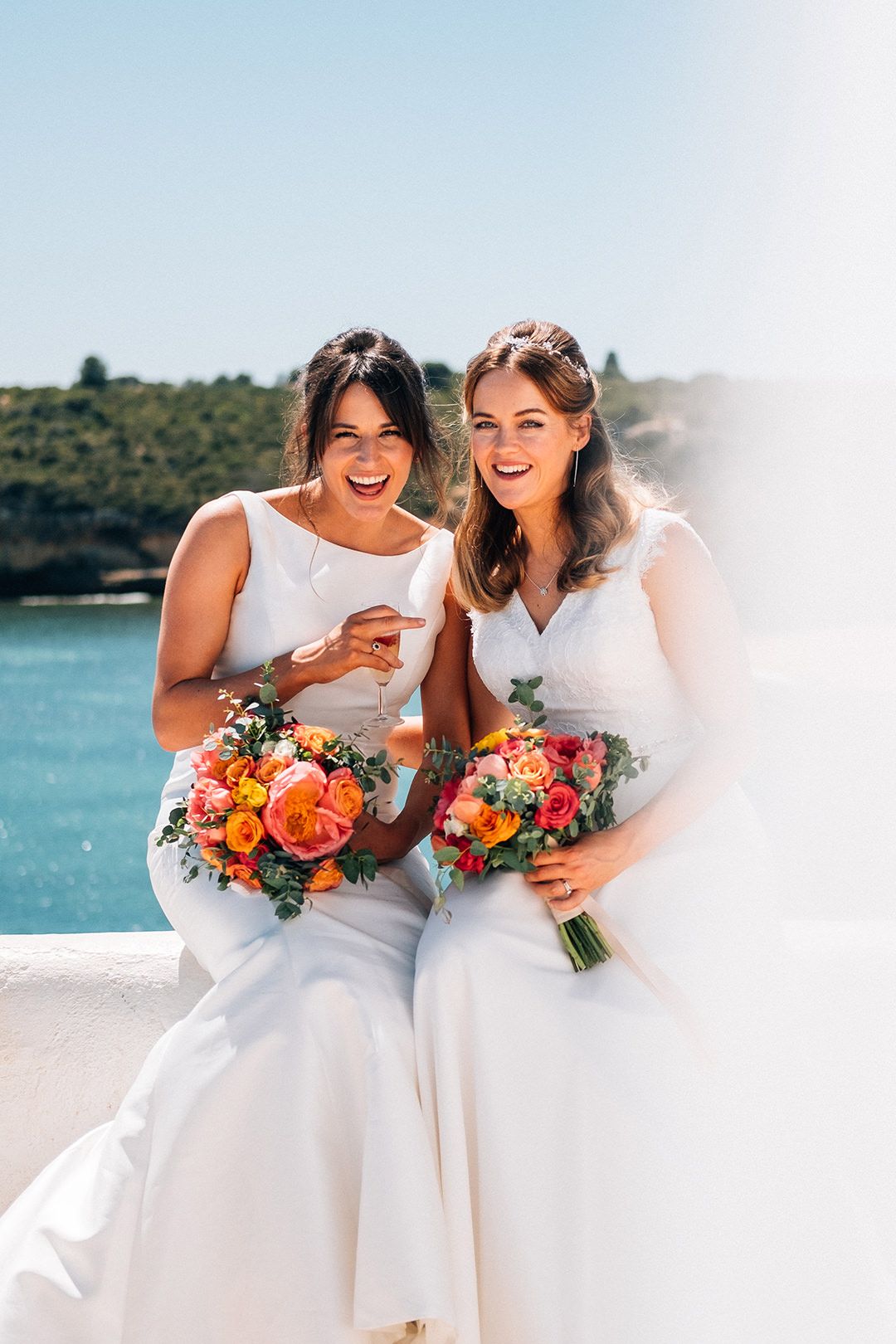 Two people in white wedding dresses share a joyful moment while holding vibrant orange and pink bouquets by the ocean.