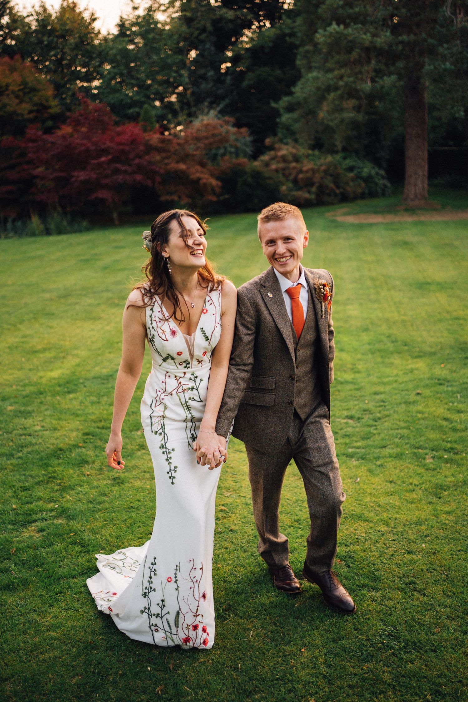Happy couple walks hand-in-hand on lush green lawn, bride in floral white gown, groom in brown tweed suit with orange tie.