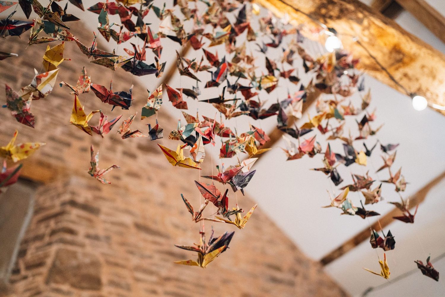 Colorful origami paper cranes hanging from strings create a delicate art installation against a textured wall.
