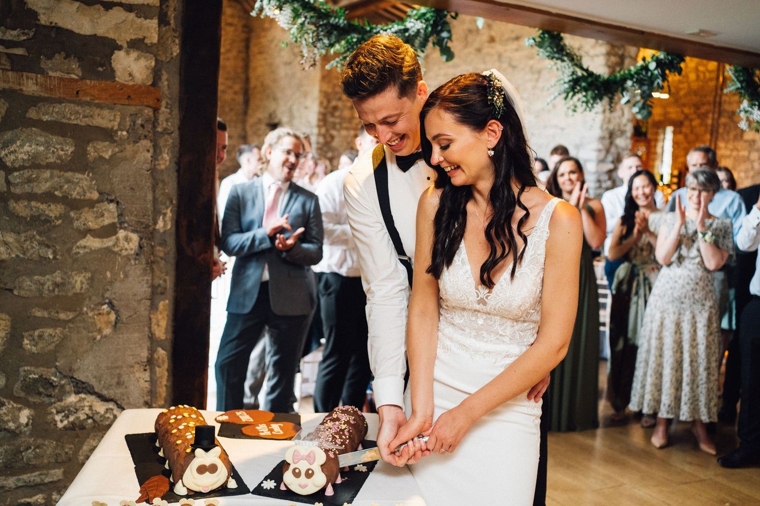 Wedding celebration with guests gathered in a historic stone barn venue decorated with greenery and white chairs.