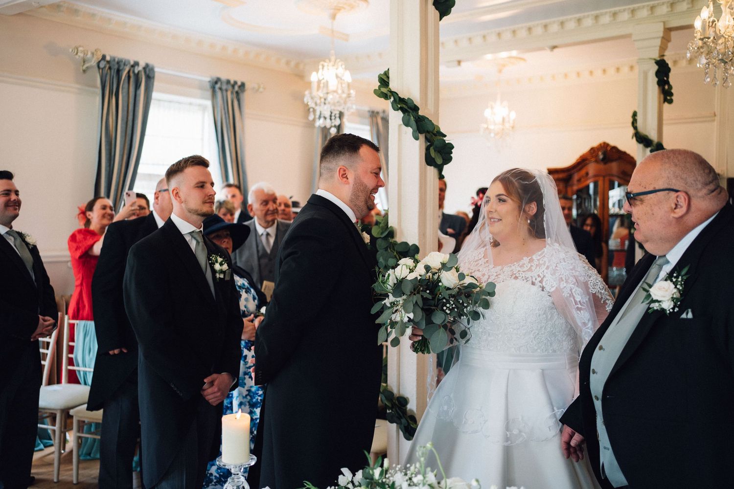 Elegant wedding ceremony in a luxurious white room with crystal chandeliers and guests seated in formal attire.