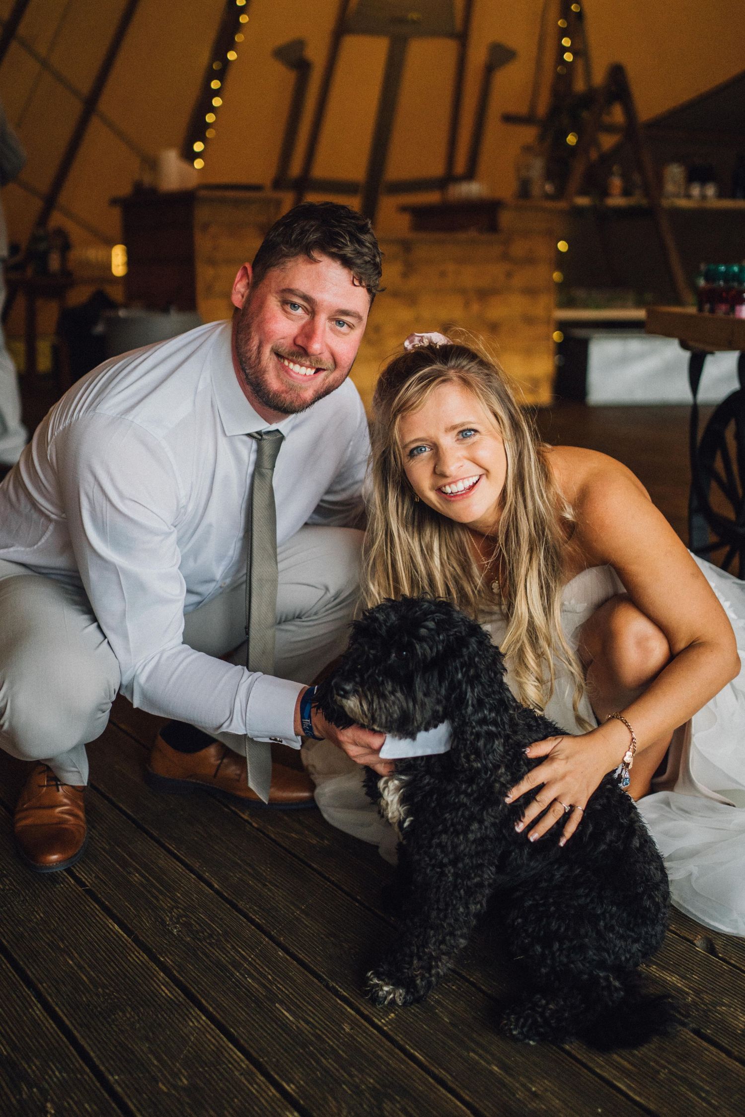 A couple poses playfully with their black dog on wooden benches under string lights.